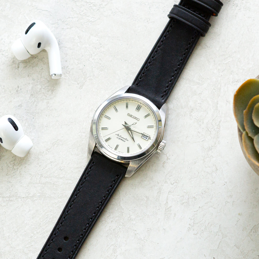 Black wristwatch with silver case on a light gray surface, next to white earbuds and a small green plant. #color_black