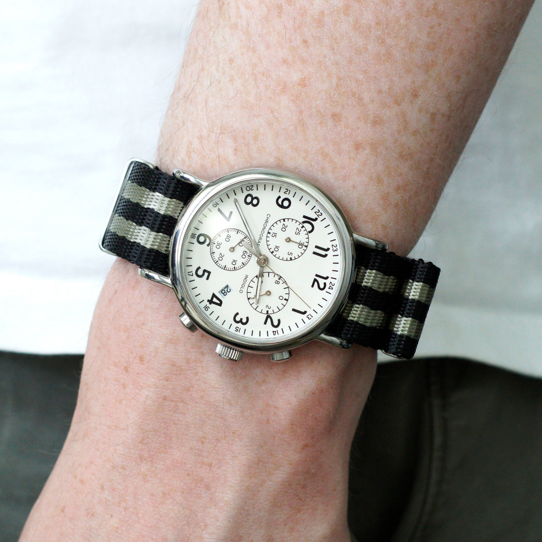 Wrist wearing a watch with a black and white striped strap on a neutral background #color_black-and-grey-regimental-striped
