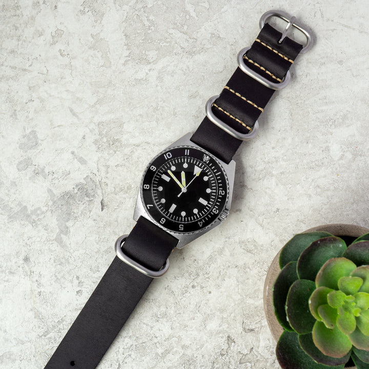 Watch with black strap and silver case on a light gray surface with a small plant. #color_black