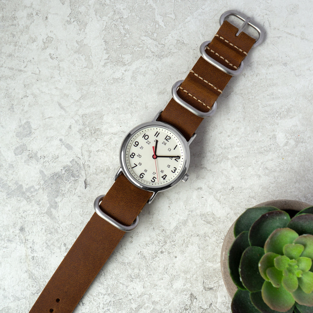 Watch with a brown strap on a light gray surface with a small plant. #color_dark-brown