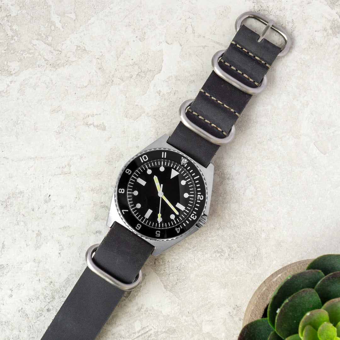 Watch with a grey strap on a light gray surface with a small plant. #color_grey