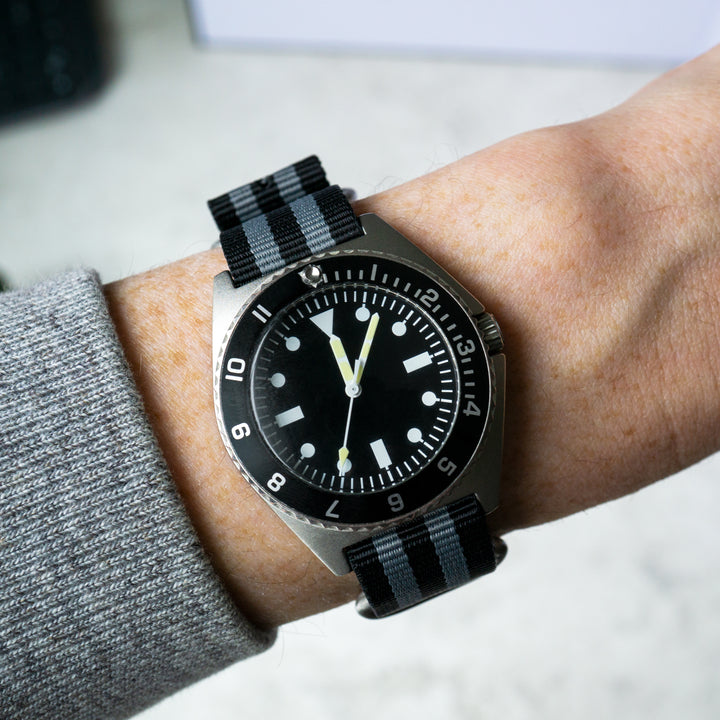 Wrist wearing a watch with a black strap and black dial on a light background #color_black-and-grey-regimental-striped
