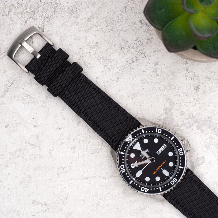 Black watch with a silver buckle on a light gray surface with a plant in the corner. #color_black