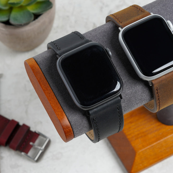 Two smartwatches with different bands on a wooden stand with a plant in the background. #color_black-pvd