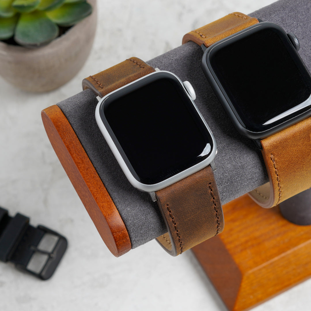 Two smartwatches with brown leather straps on a wooden stand with a plant in the background. #color_dark-brown