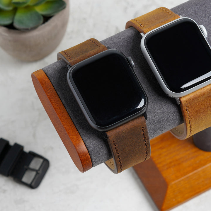 Two smartwatches with brown leather straps on a wooden stand, with a plant in the background. #color_dark-brown-pvd