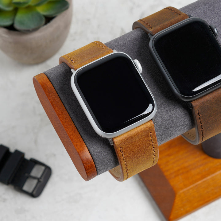 Two smartwatches with brown leather straps on a wooden stand with a plant in the background. #color_sand