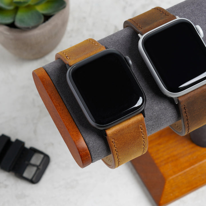 Two smartwatches with brown leather straps on a wooden stand, with a small plant in the background. #color_sand-pvd
