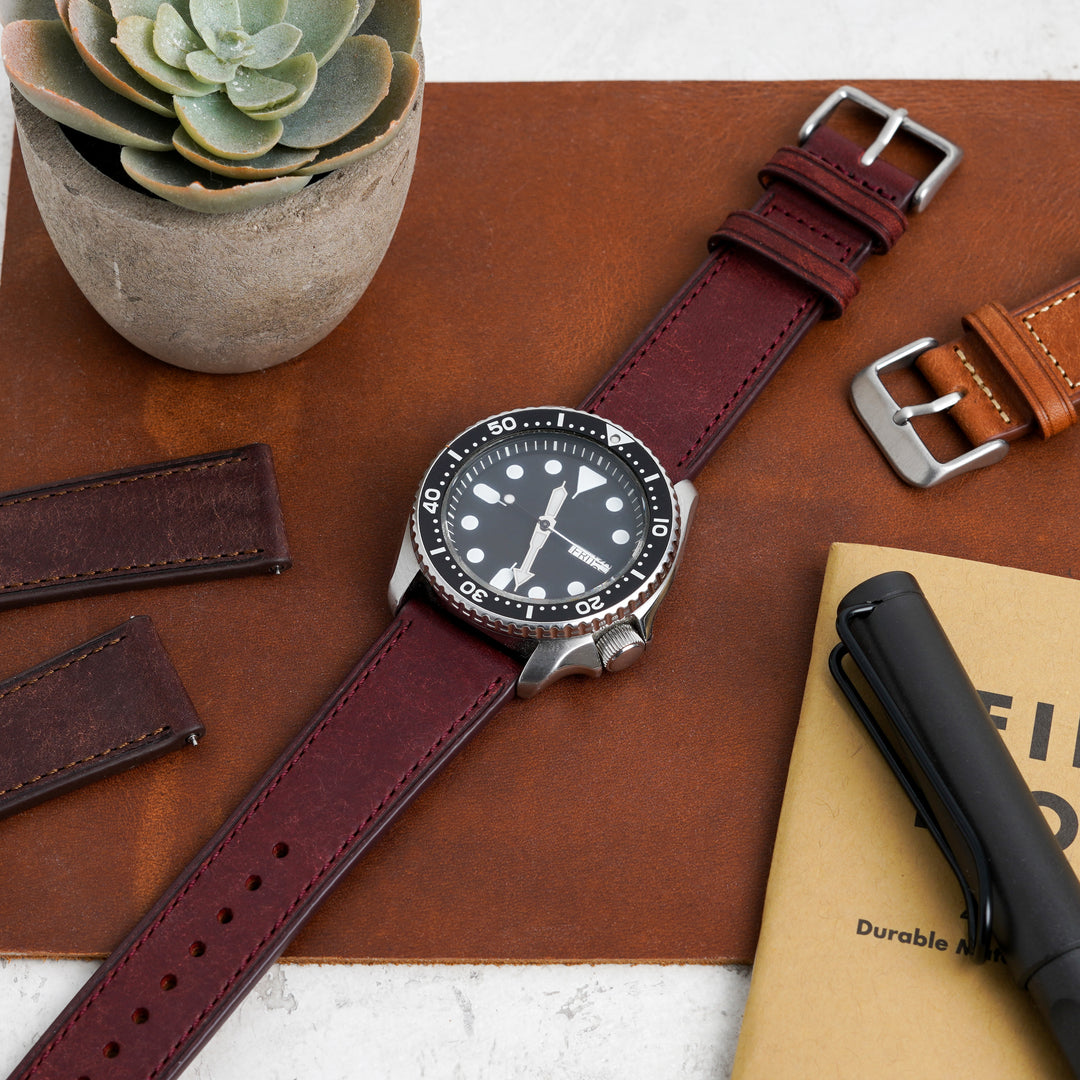 Watch with brown leather strap on a wooden surface with a plant and notebook. #color_burgundy