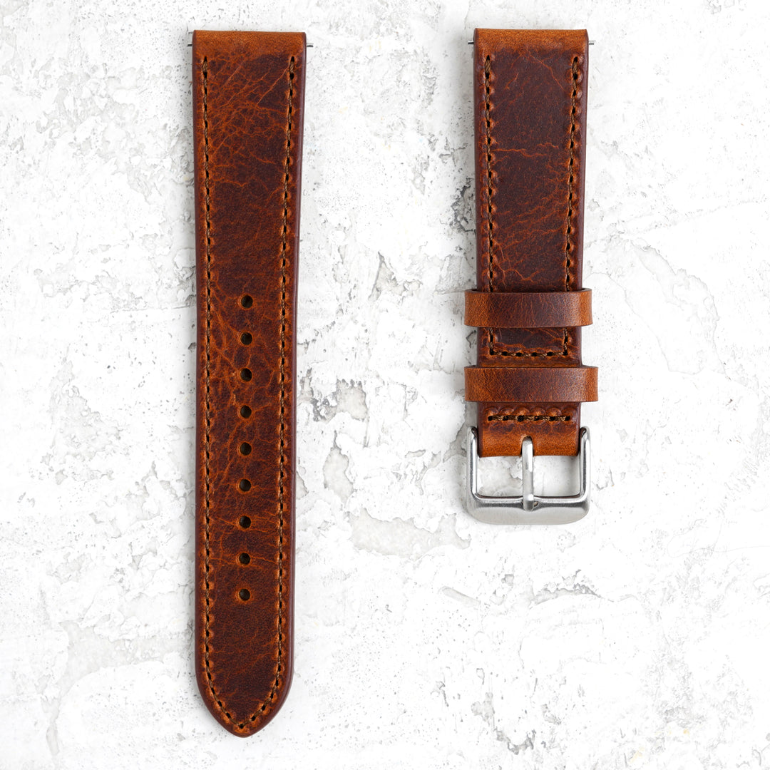A brown waxy vegetable-tanned leather watch band with a brushed stainless-steel buckle and stitching details, displayed on a textured white background. #color_rust